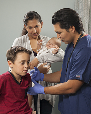 Un proveedor de atención médica le pone una inyección a un niño. Una madre sostiene a su bebé en segundo plano.